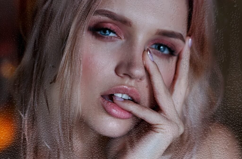 Aantrekkelijk jong wijfje met blauwe ogen roze oogschaduw en sensuele lippen die vinger bijten en gezicht met vingers aanraken en naar camera kijken, liplift