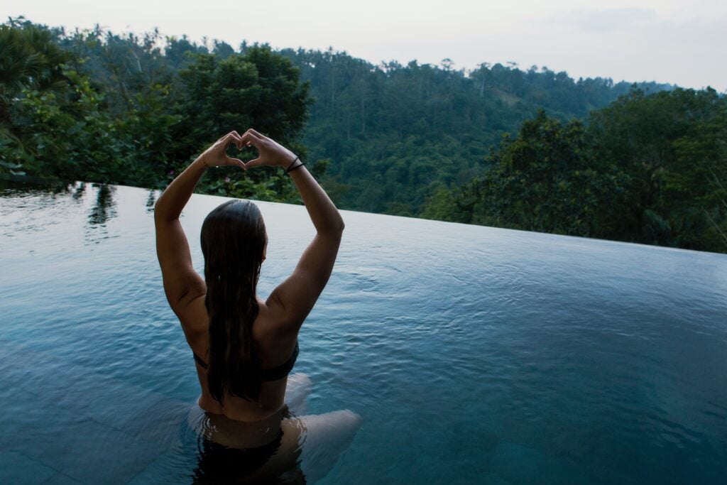 femme dans une piscine à débordement faisant un geste de la main en forme de cœur face à des arbres à feuilles vertes, chirurgie de féminisation du corps